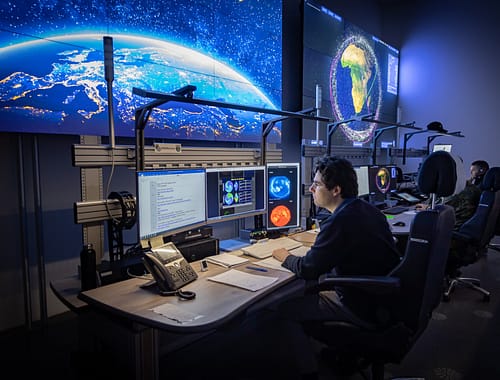 A control room operator looks at satellite imagery from a KVM station connected via VuWall’s video wall management system. The workspace features monitors, a phone, and paperwork from the German Space Agency.