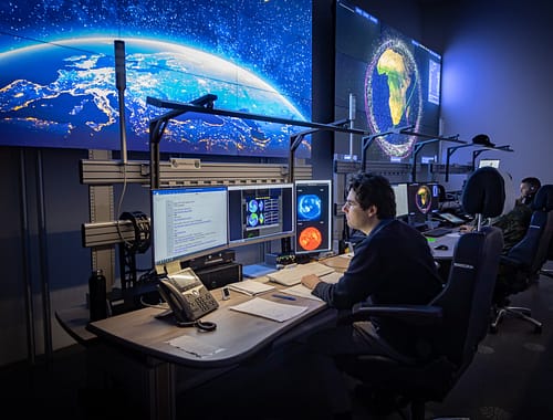 Un operador de la sala de control observa imágenes de satélite desde una estación KVM conectada mediante el sistema de gestión de videowall de VuWall. En el espacio de trabajo hay monitores, un teléfono y documentación de la Agencia Espacial Alemana.