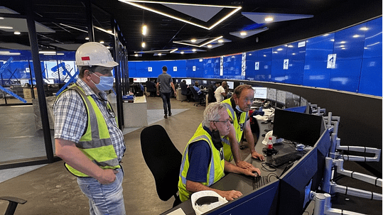 Tres hombres con chalecos y cascos de seguridad trabajan en una sala de control de minería de vanguardia que cuenta con soluciones avanzadas de videowalls de VuWalls. Dos ingenieros observan mientras un colega maneja un ordenador, optimizando las operaciones. Las grandes pantallas azules de las paredes muestran el elegante diseño 2025 de VuWalls, que ofrece una integración perfecta y una mejor toma de decisiones para los profesionales audiovisuales y los integradores de sistemas.