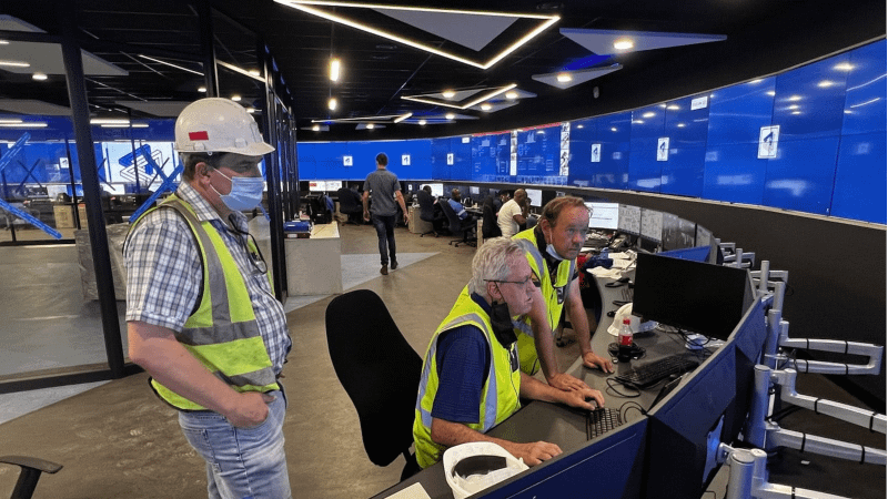 Three men in safety vests and hard hats work in a cutting-edge mining control room featuring VuWalls advanced video wall solutions. Two engineers observe while a colleague operates a computer, optimizing operations. Large blue screens on the walls showcase VuWalls sleek 2025 design, offering seamless integration and enhanced decision-making for AV professionals and system integrators.